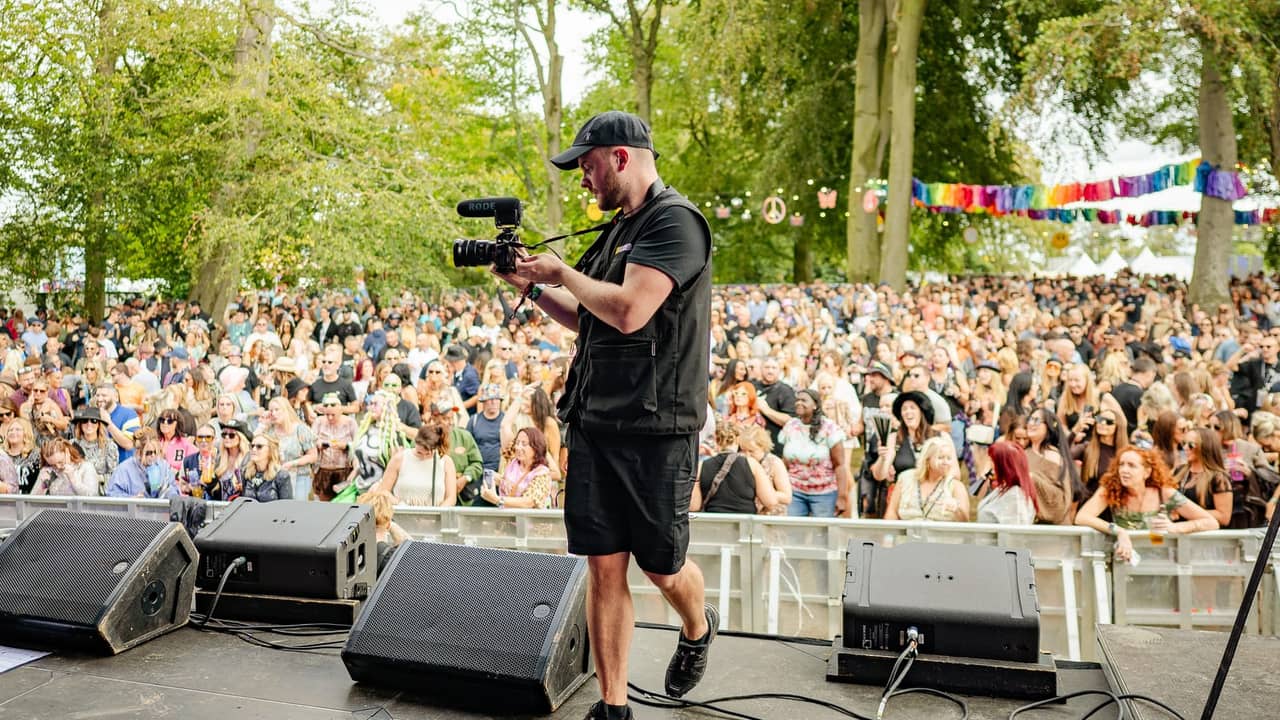 A videographer stands on a festival stage holding a mirrorless camera with a shotgun microphone, filming the large crowd gathered beyond the barrier under trees strung with rainbow bunting.