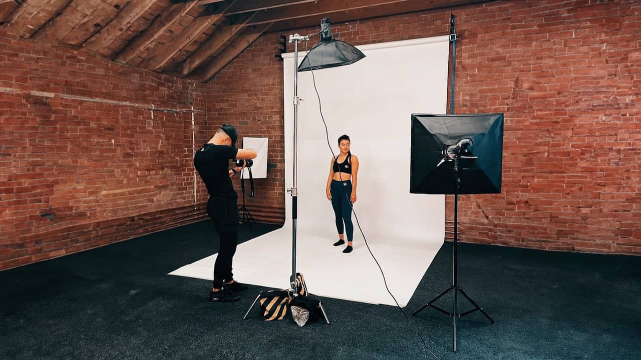 Photographer shooting a model in sportswear against white backdrop with softbox and umbrella lighting.