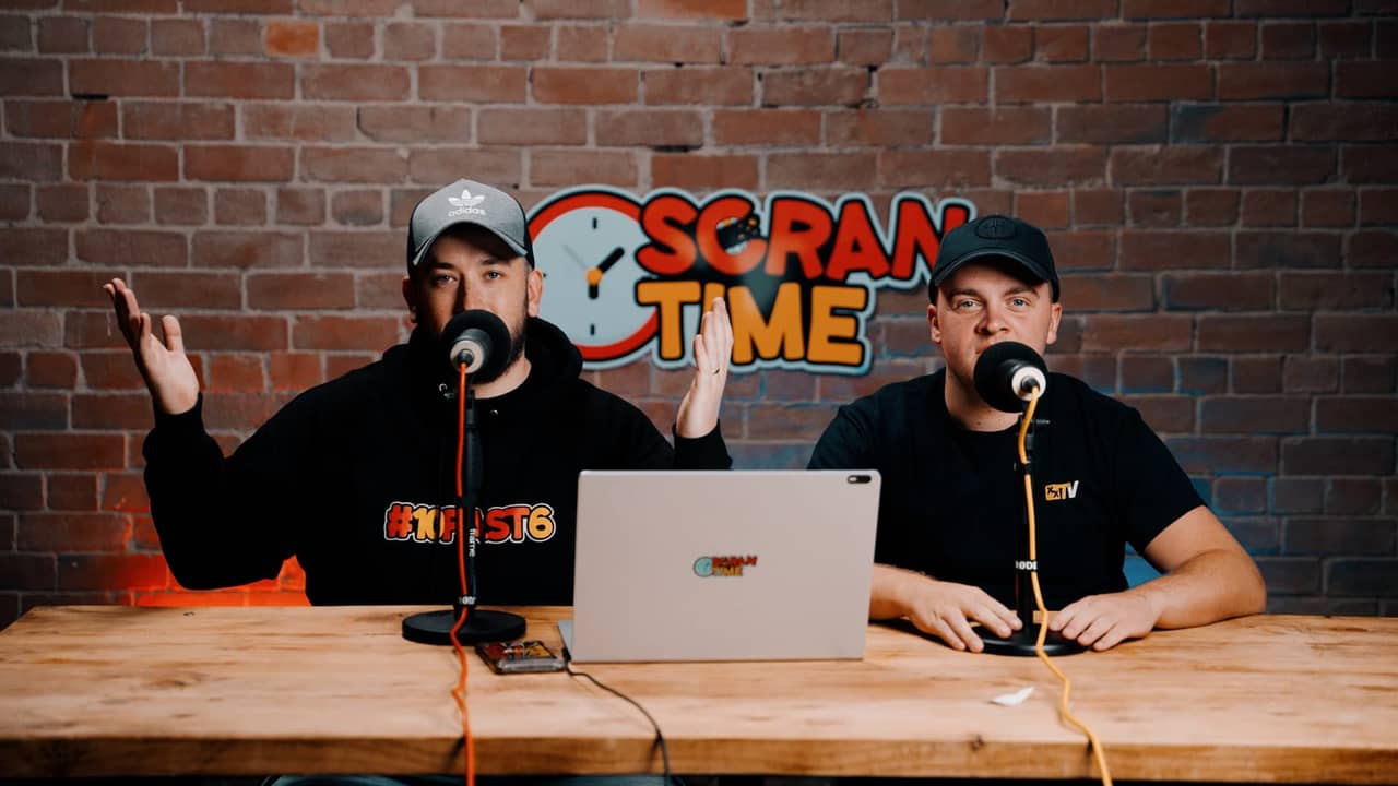 Two podcast hosts sit at a wooden table with microphones and a laptop in front of a brick wall branded with the show's logo, one gesturing while speaking.