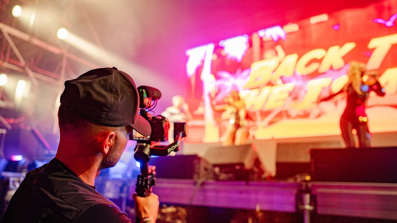 Camera operator using a gimbal rig to film performers on a brightly lit festival stage