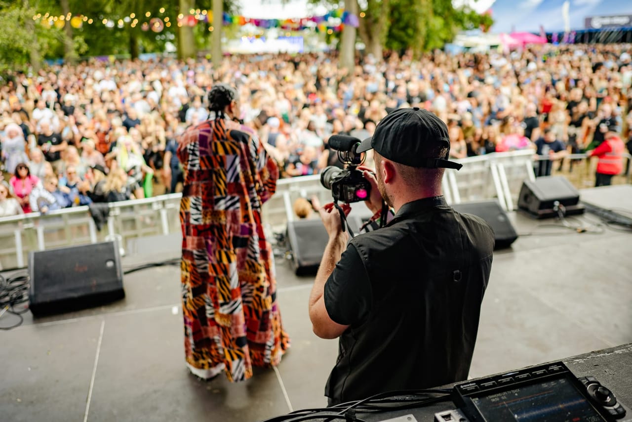 A Flexxed camera operator films a performer in a bold patterned robe from behind on a festival stage, facing a packed crowd