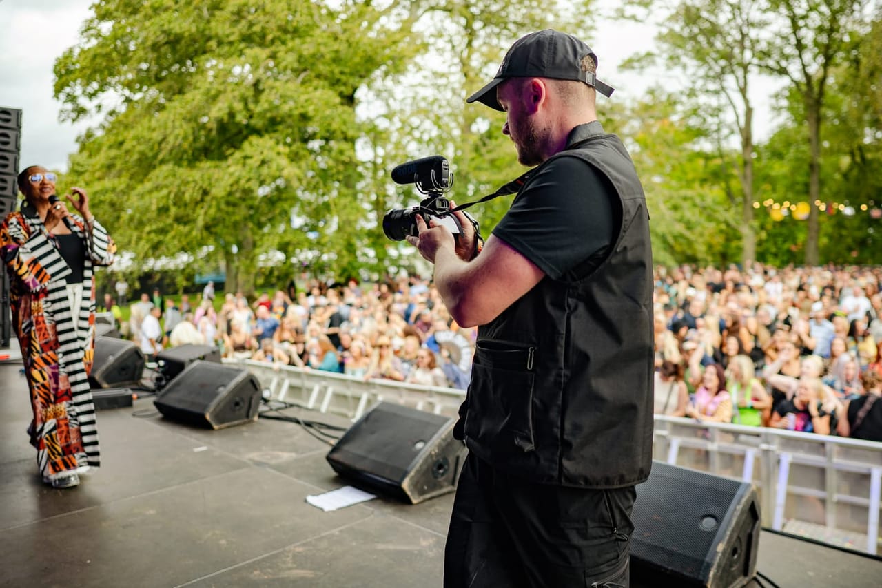 A camera operator in a Flexxed vest films a performer from the side of a festival stage, the crowd and trees behind