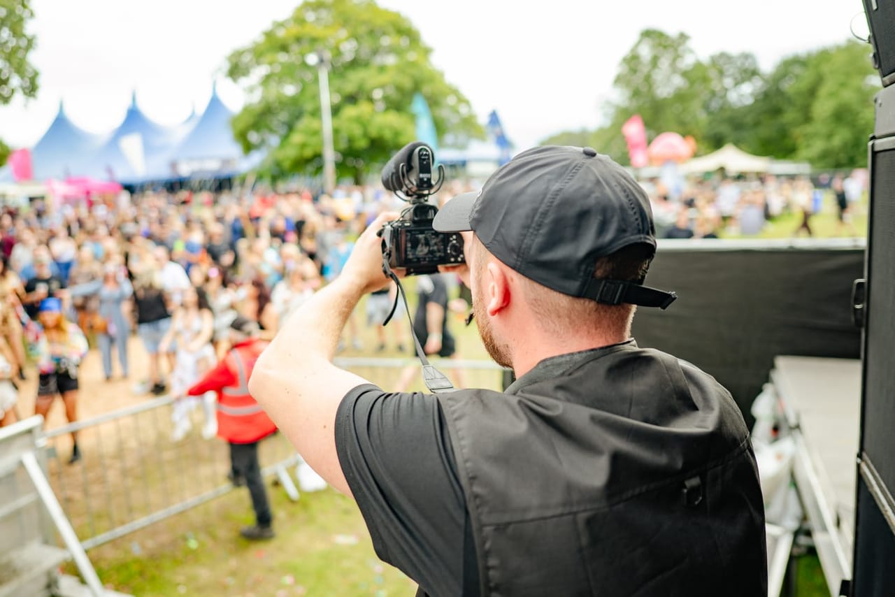 A camera operator leans over the front of the stage to film the festival crowd, big-top tents in the distance