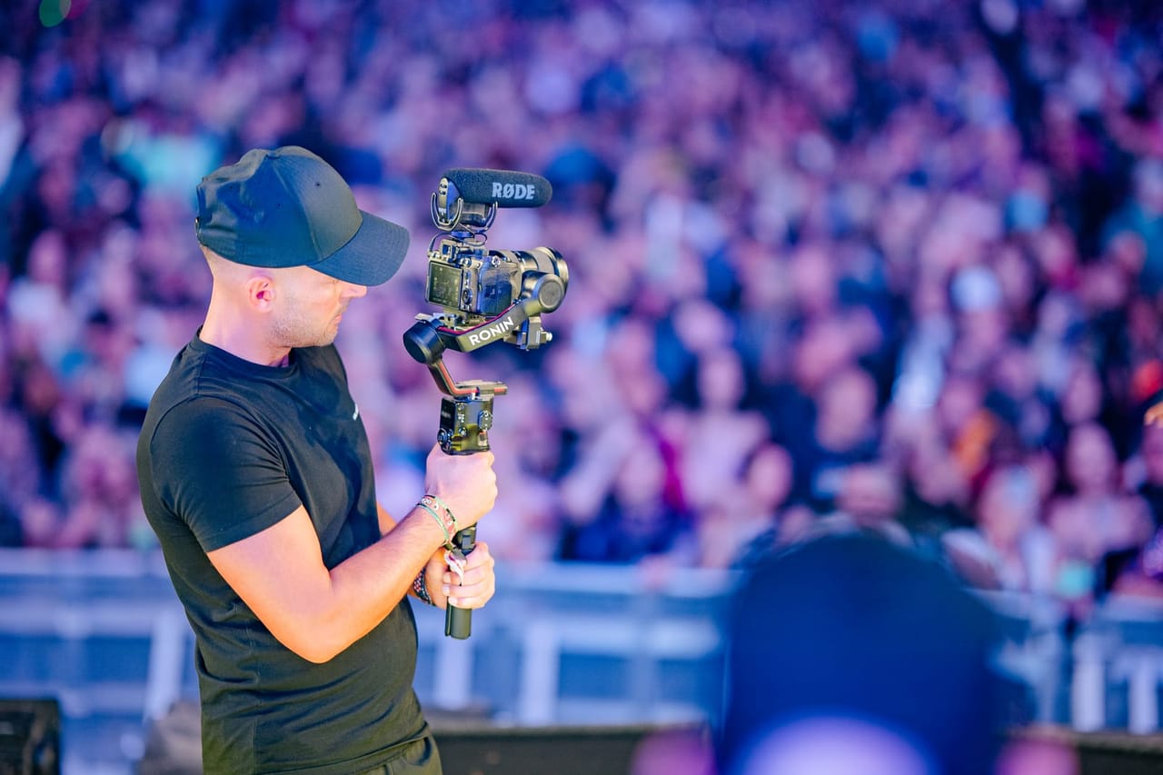A camera operator in a cap works his gimbal rig in front of a packed crowd under purple stage light
