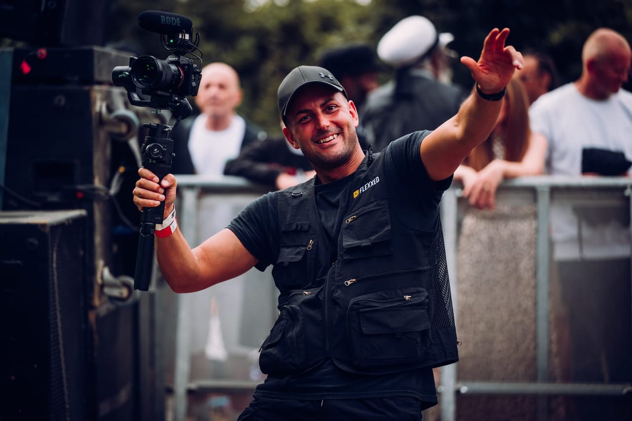 A smiling Flexxed camera operator waves to the crowd at the barrier with a gimbal-mounted camera in his other hand
