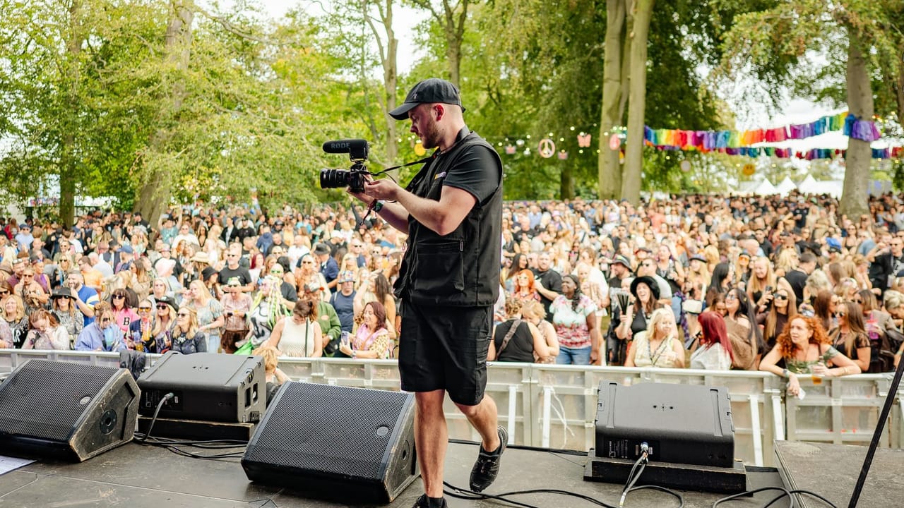 A videographer stands on a festival stage holding a mirrorless camera with a shotgun microphone, filming the large crowd gathered beyond the barrier under trees strung with rainbow bunting.