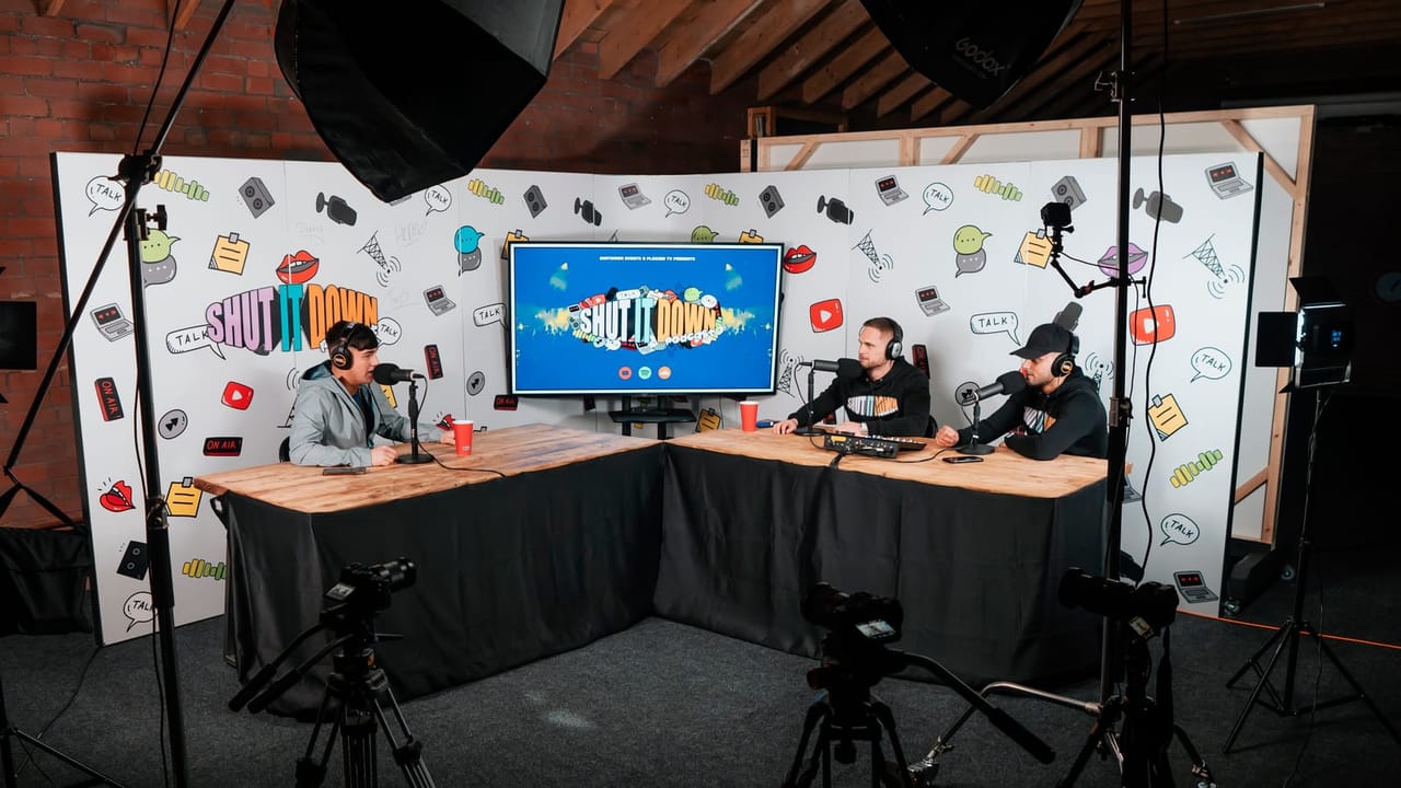 Three hosts on headphones and microphones at the Shut It Down podcast desk in the Darwen studio, cameras and softbox lights set around them