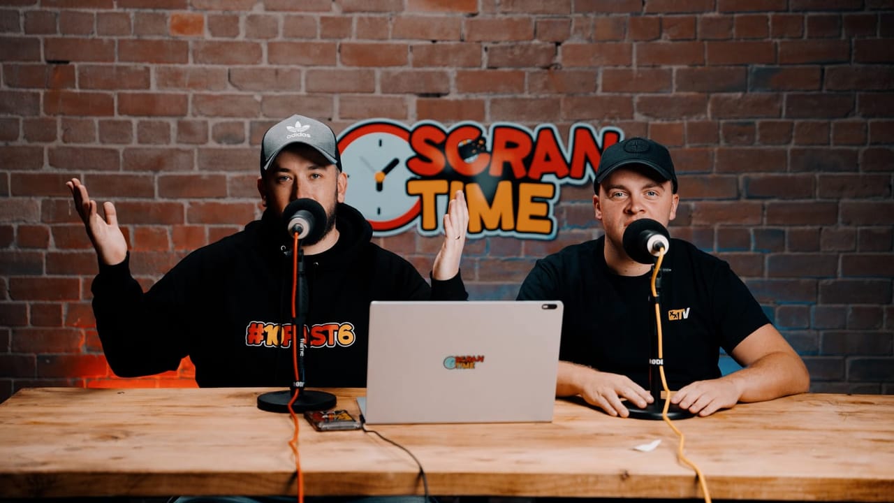 Two podcast hosts sit at a wooden table with microphones and a laptop in front of a brick wall branded with the show's logo, one gesturing while speaking.