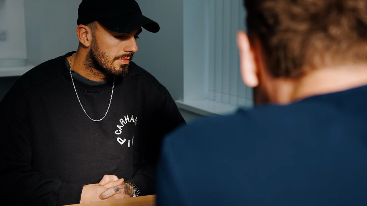 Man in a cap being interviewed across a desk in a clinic consultation room, with the interviewer's shoulder in the foreground