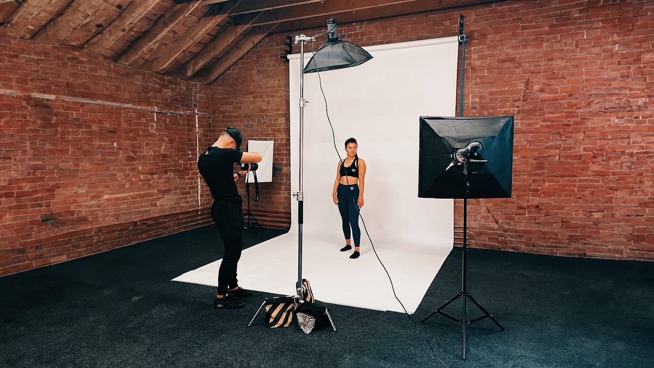 Photographer shooting a model in sportswear against white backdrop with softbox and umbrella lighting.