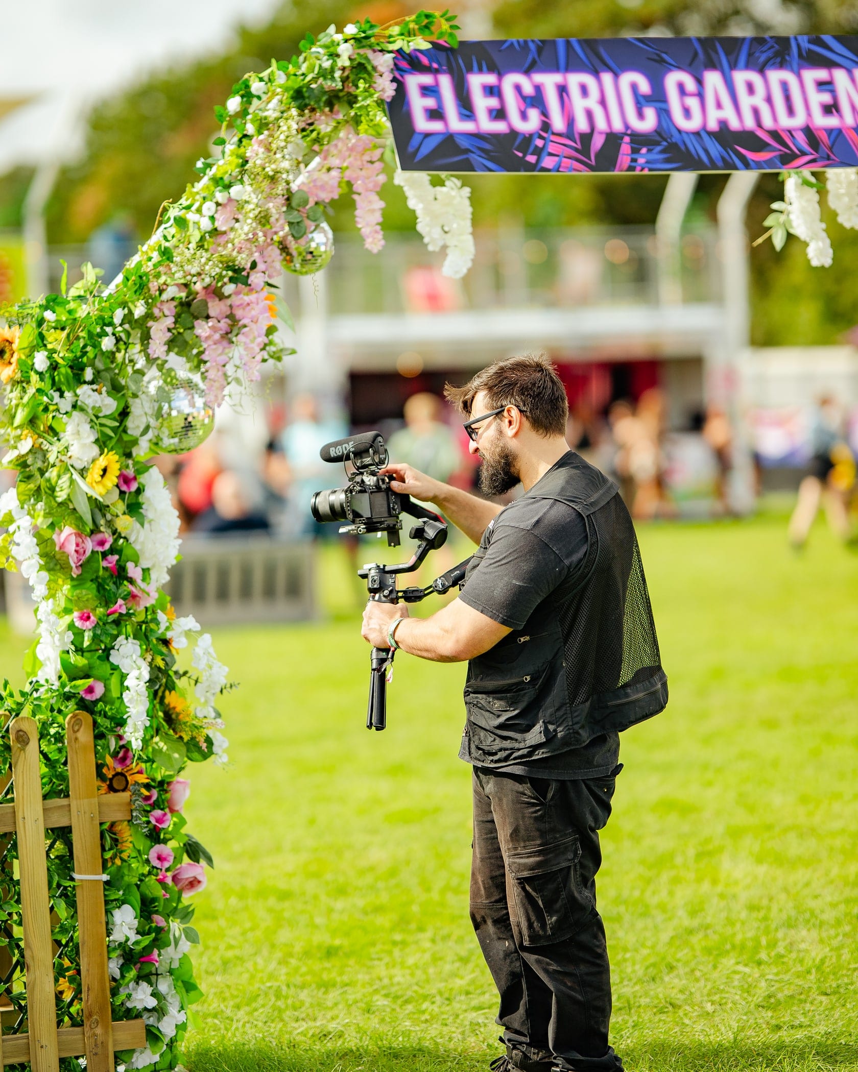 A Flexxed camera operator setting up a gimbal shot under the flower-covered Electric Garden arch at a festival