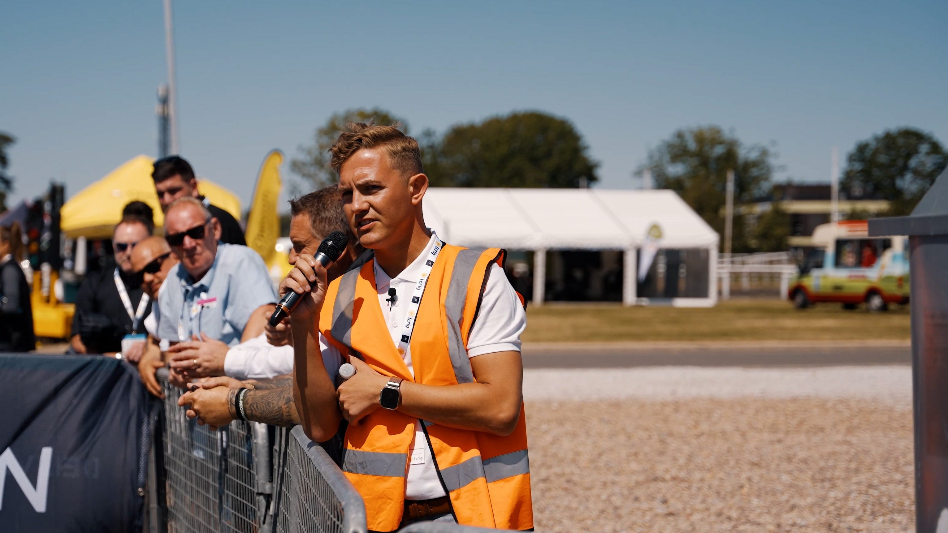 A presenter in a hi-vis vest on a handheld microphone at a trade demonstration, visitors along the barrier behind him