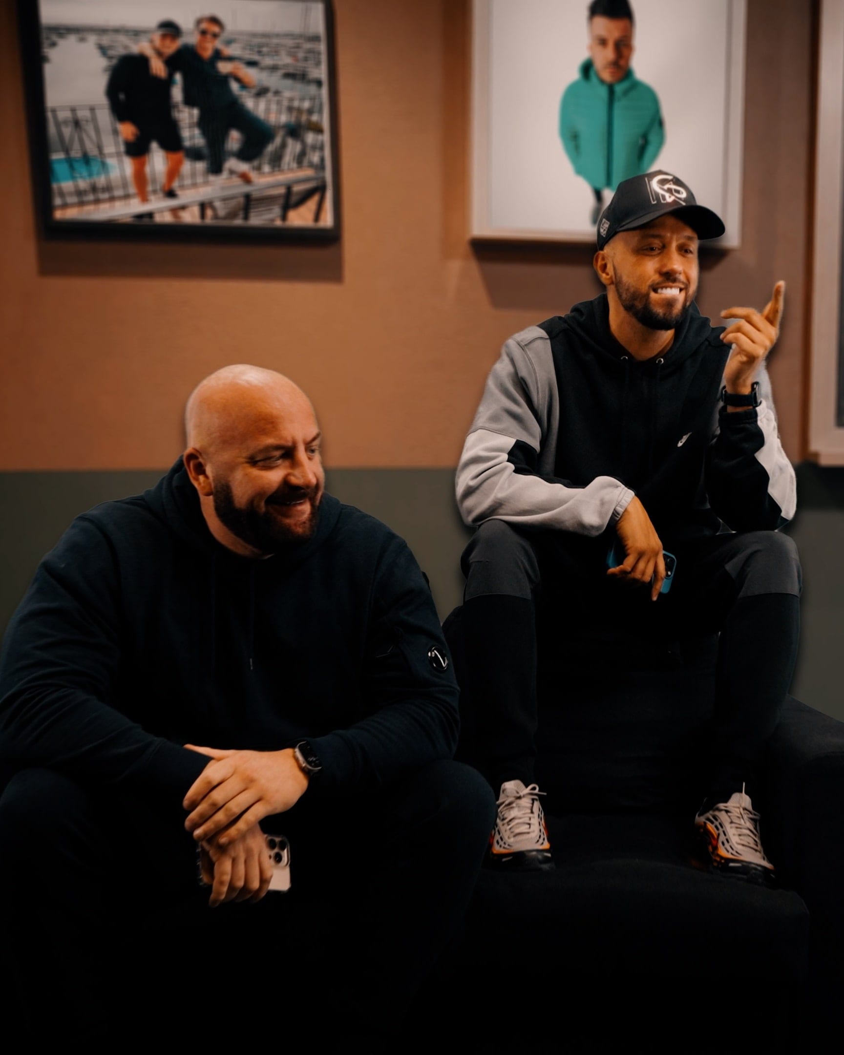 Two artists on a sofa in a lounge set between takes, framed photographs on the wall behind them