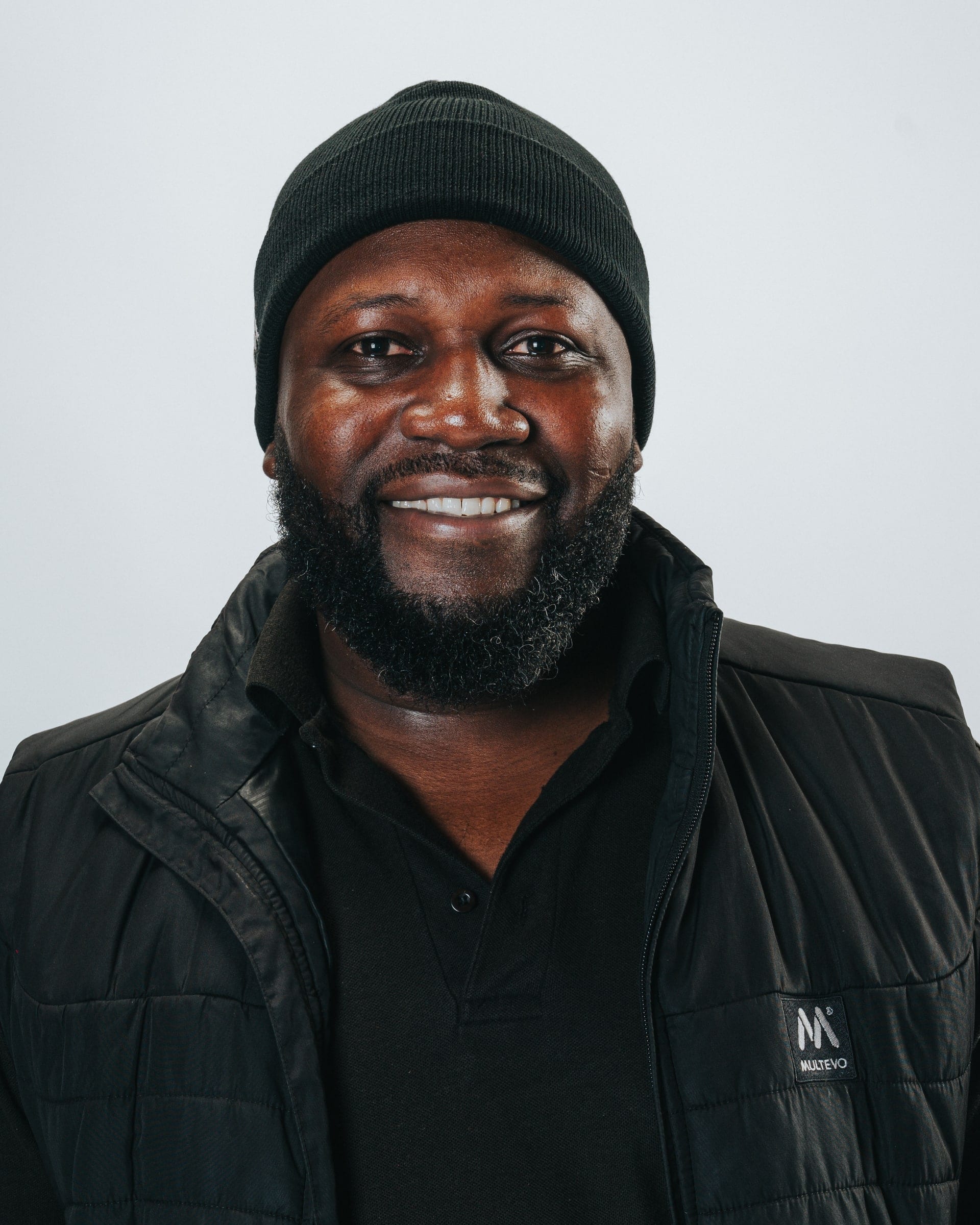 Corporate headshot of a smiling man in a black beanie and black jacket, on a pale grey backdrop