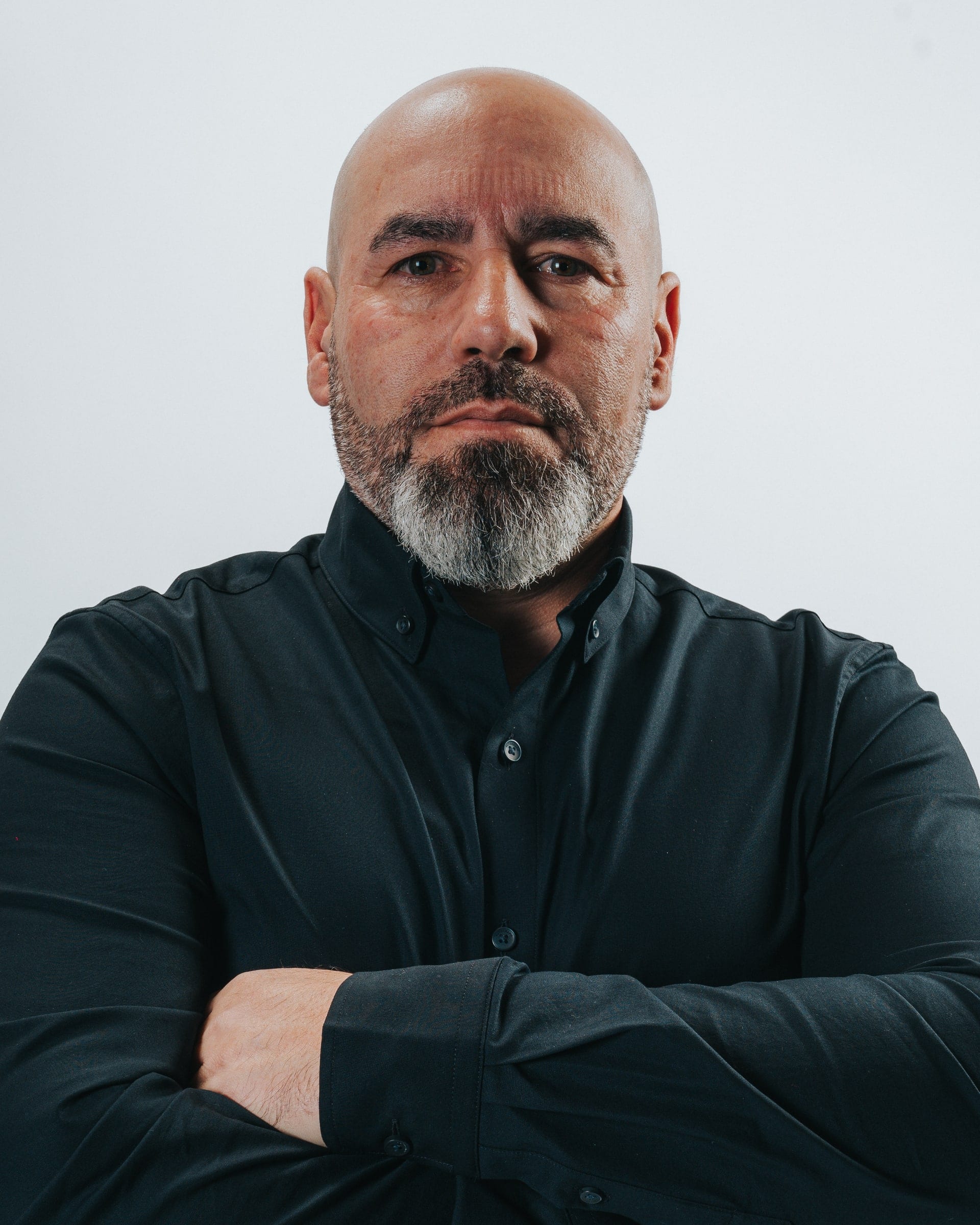 Corporate headshot of a bald man with a grey beard in a black shirt, arms folded, on a pale grey backdrop