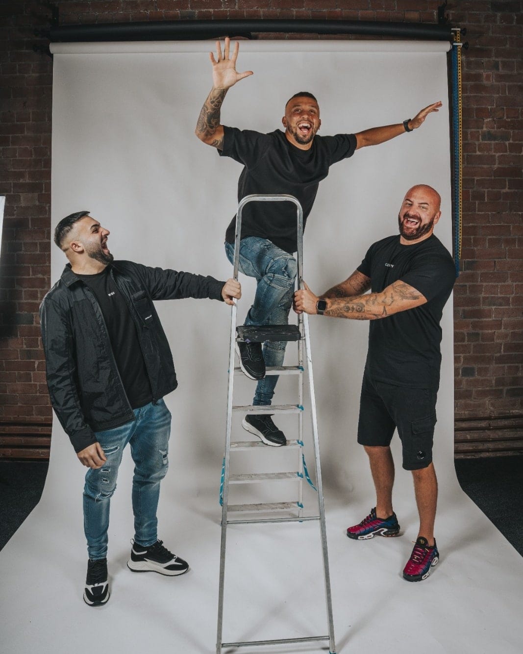 Three men laughing on a white cyclorama against a brick wall, one standing on an aluminium stepladder with his arms flung out while the other two hold it steady