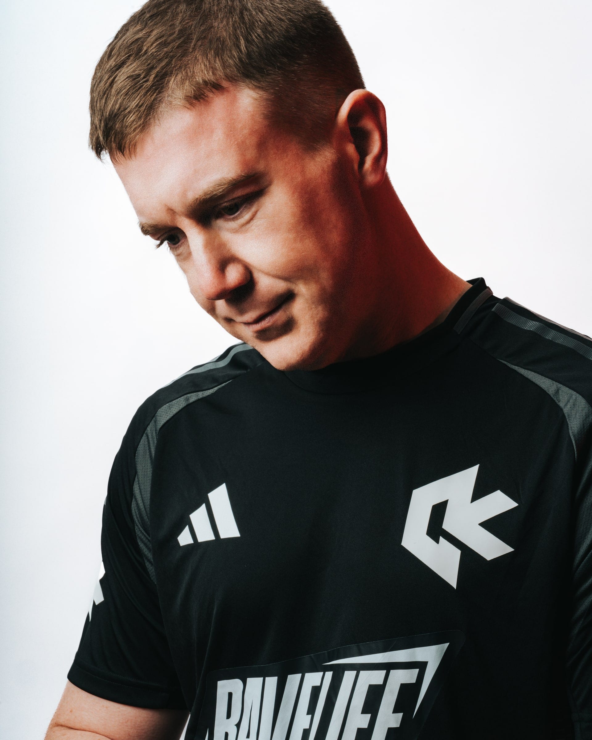 A man in a black Ravelife football shirt, head bowed, lit against a white backdrop