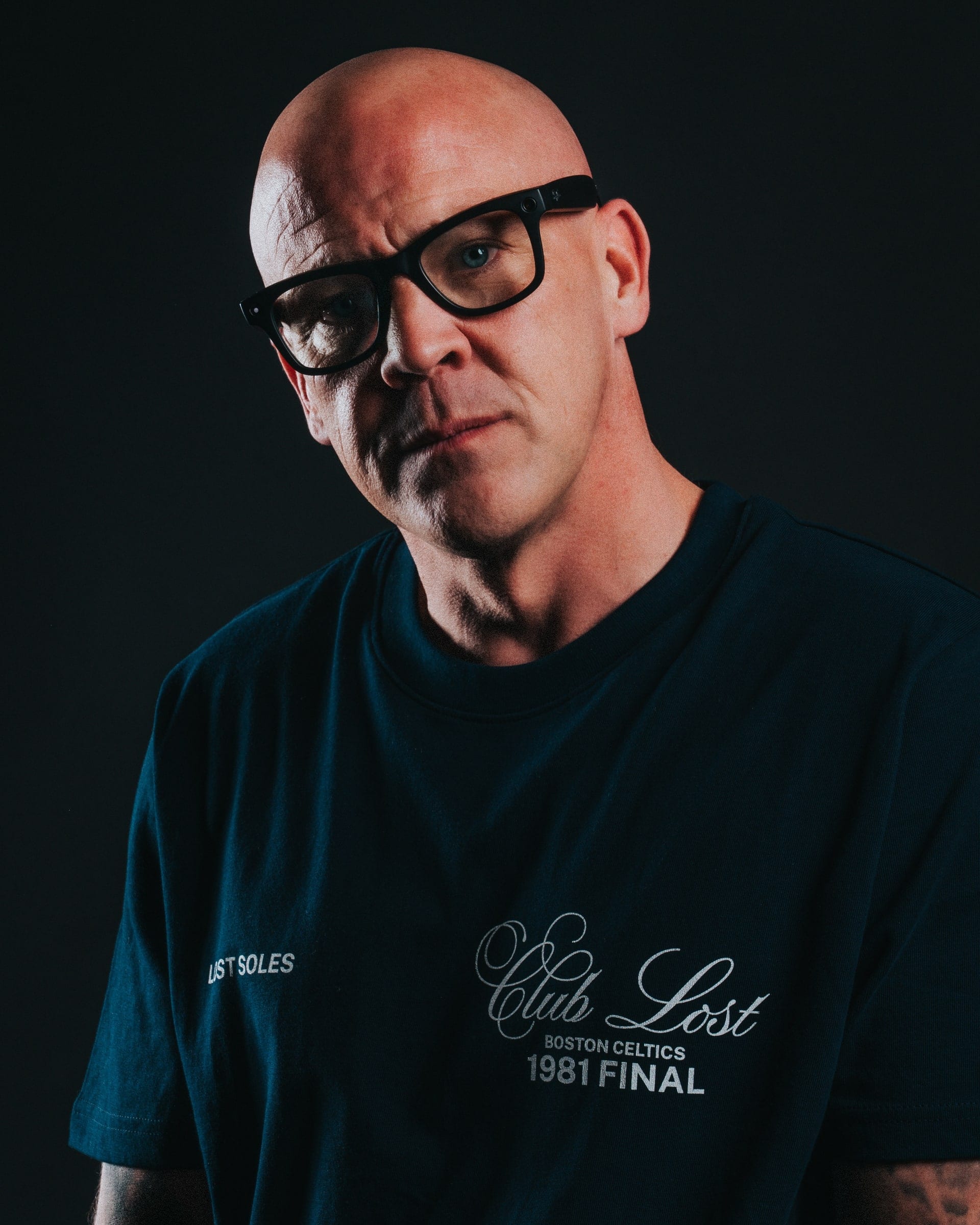 A bald man in heavy framed glasses and a navy tee, lit hard from one side