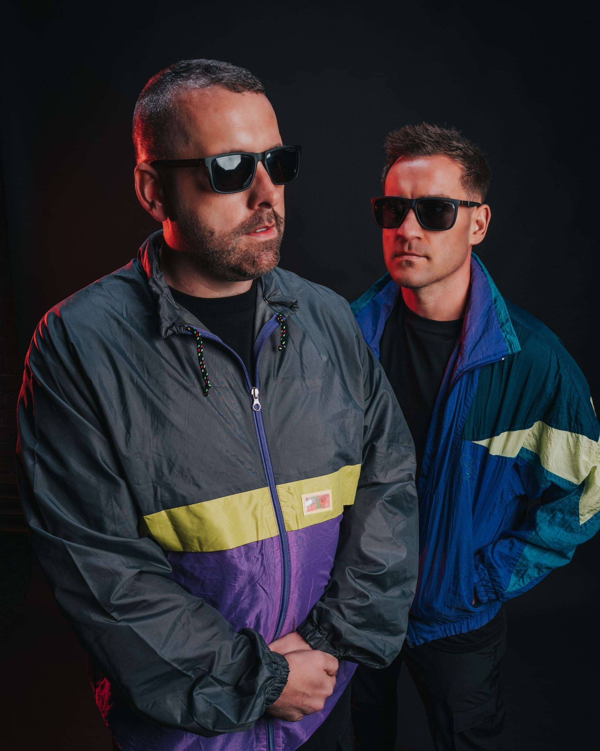 Two men in retro shell jackets and dark glasses, side by side against a dark backdrop