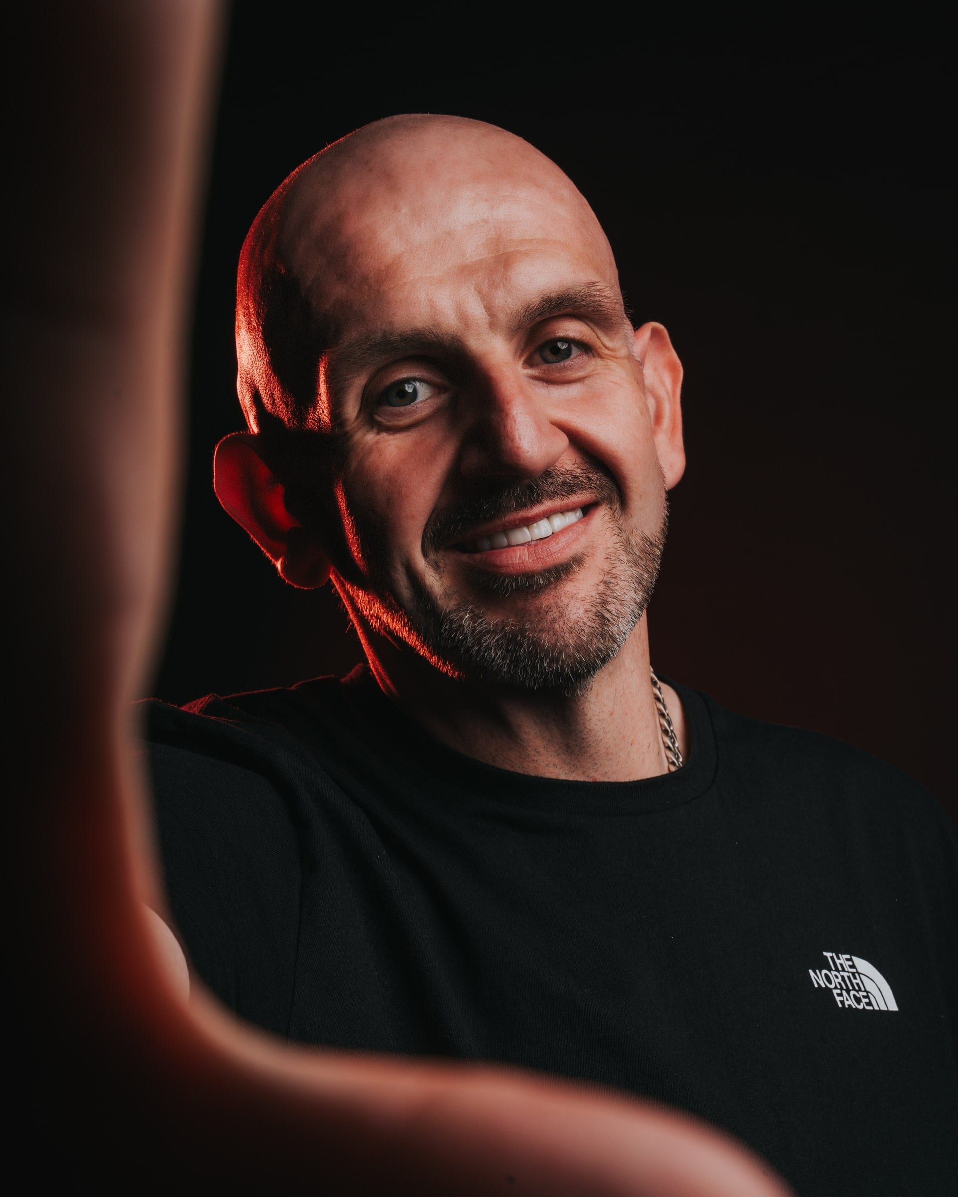 A bald man smiling in red light, an arm crossing the frame in the foreground