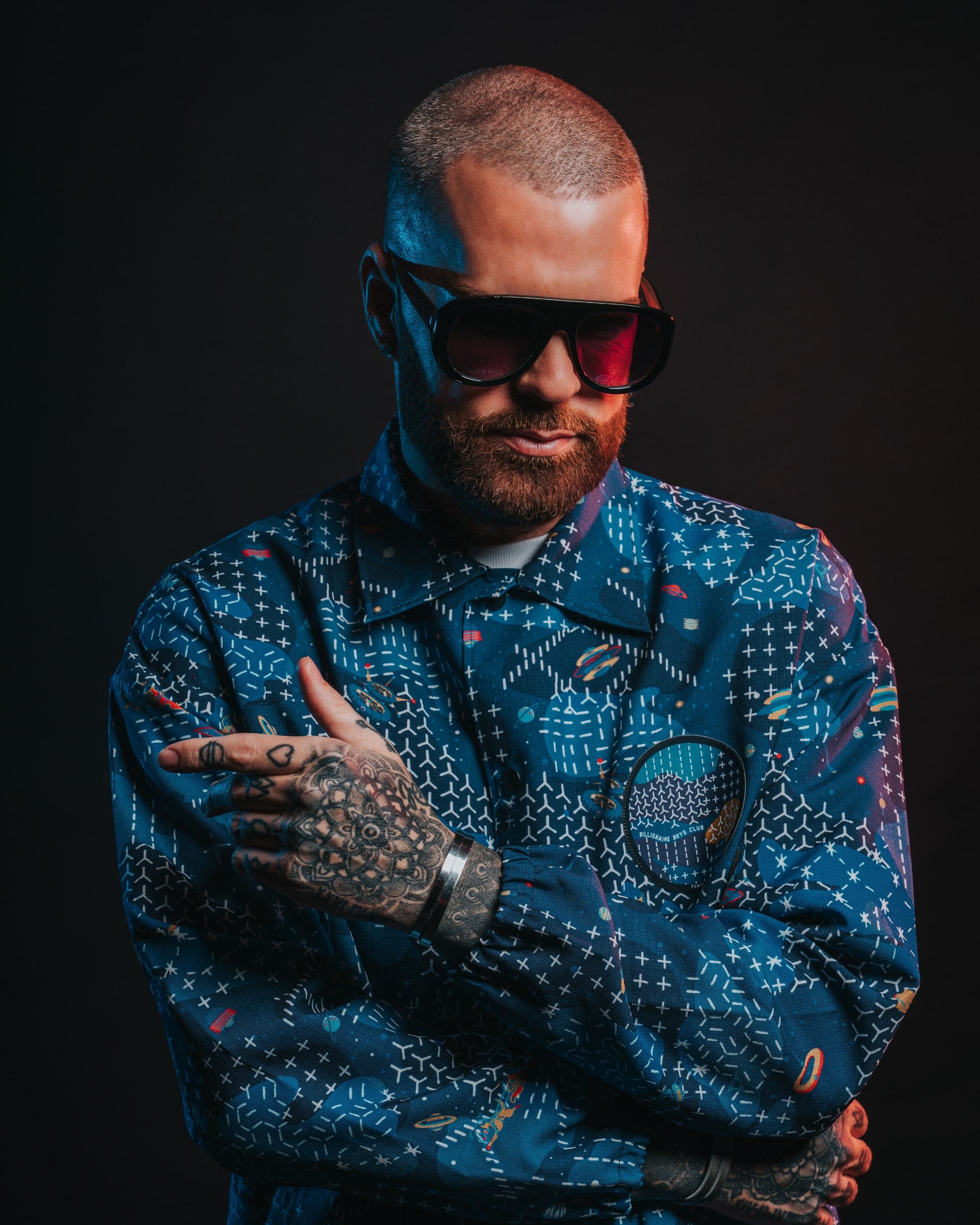 A tattooed man in dark glasses and a blue paisley shirt, arms folded