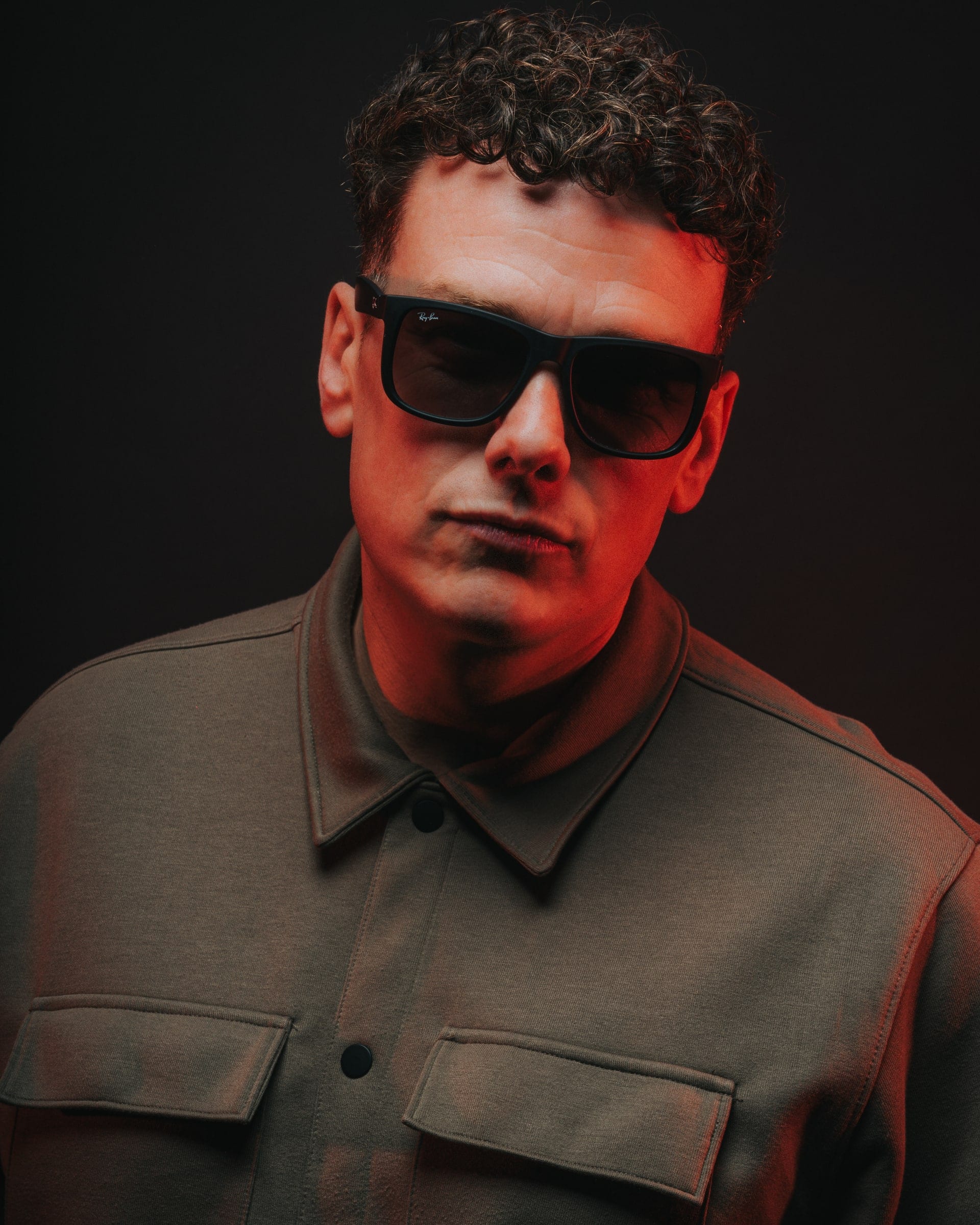 A man with curly hair in dark glasses and an olive overshirt under warm light