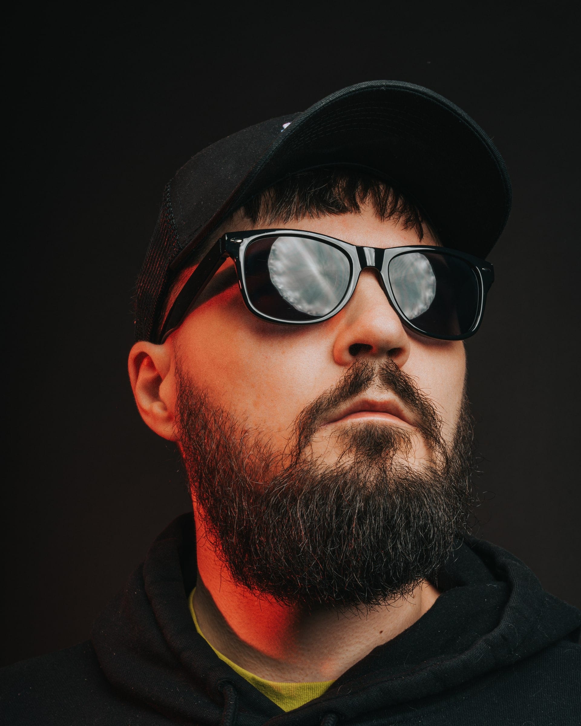 A bearded man in mirrored sunglasses and a cap, lit from below in red