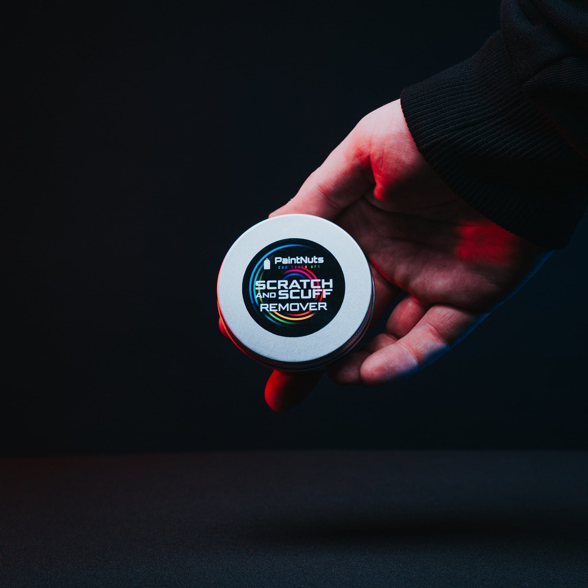 A hand holding out a PaintNuts scratch and scuff remover tin against black, rim lit in red
