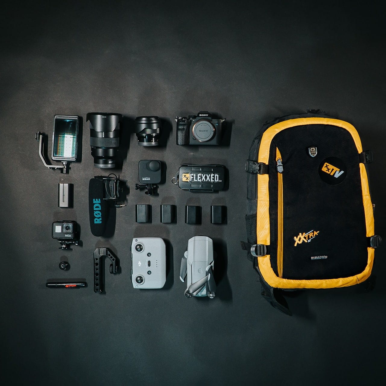 A camera kit laid out flat on black: bodies, lenses, a drone controller and a yellow backpack