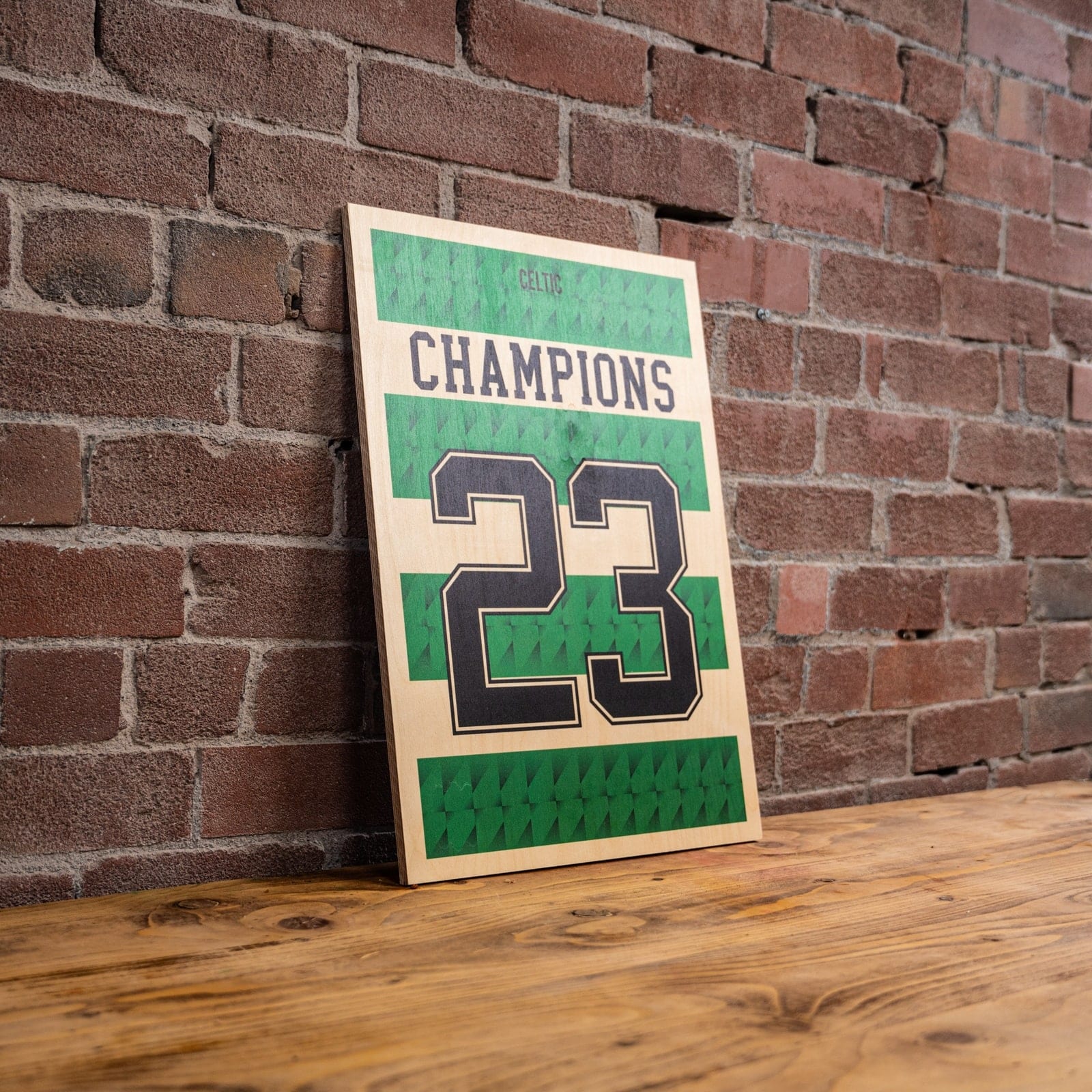 A framed green Champions 23 football shirt design leaning against a brick wall