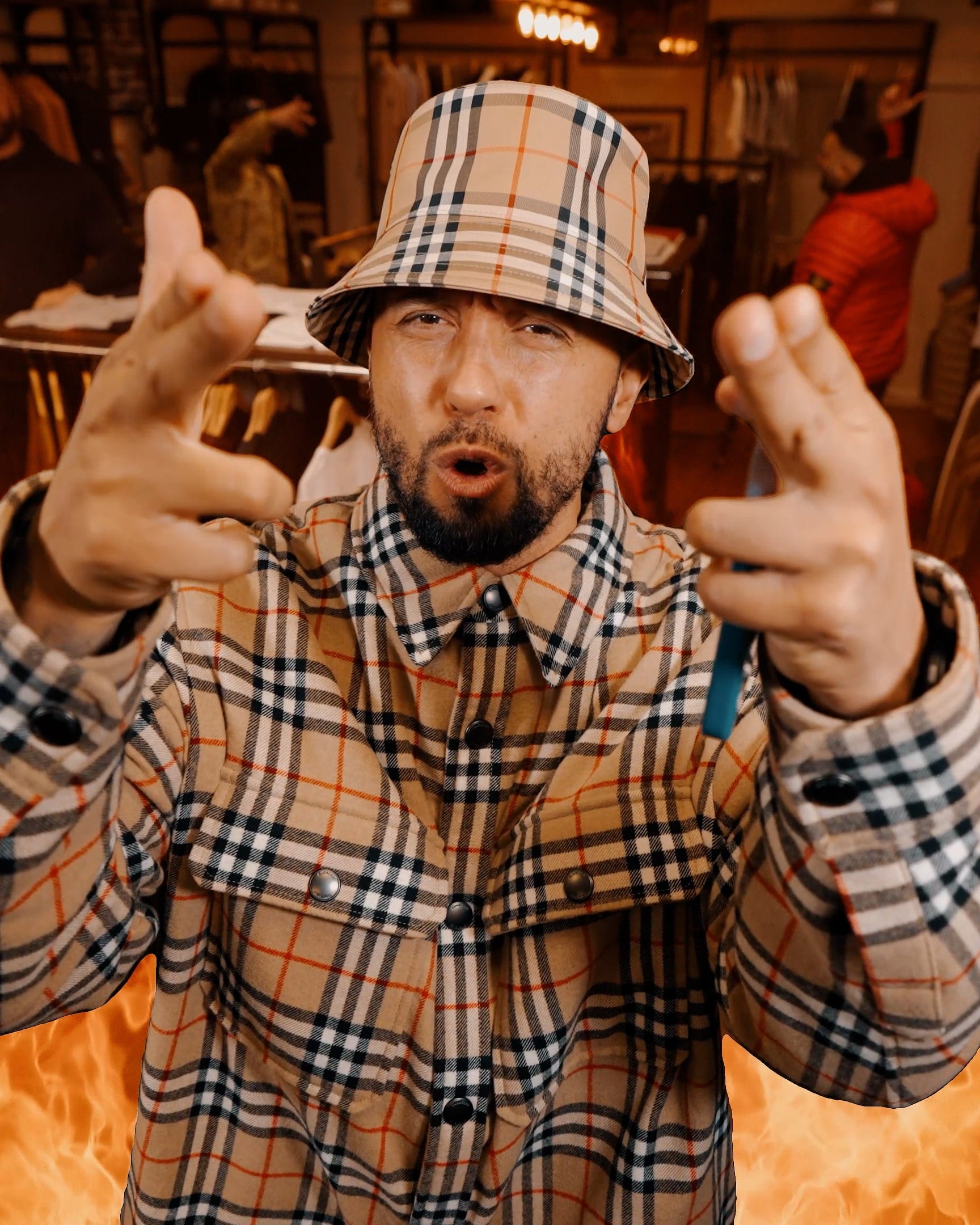 A rapper in a checked bucket hat and matching jacket, hands raised to camera, flames burning behind him on a music video set
