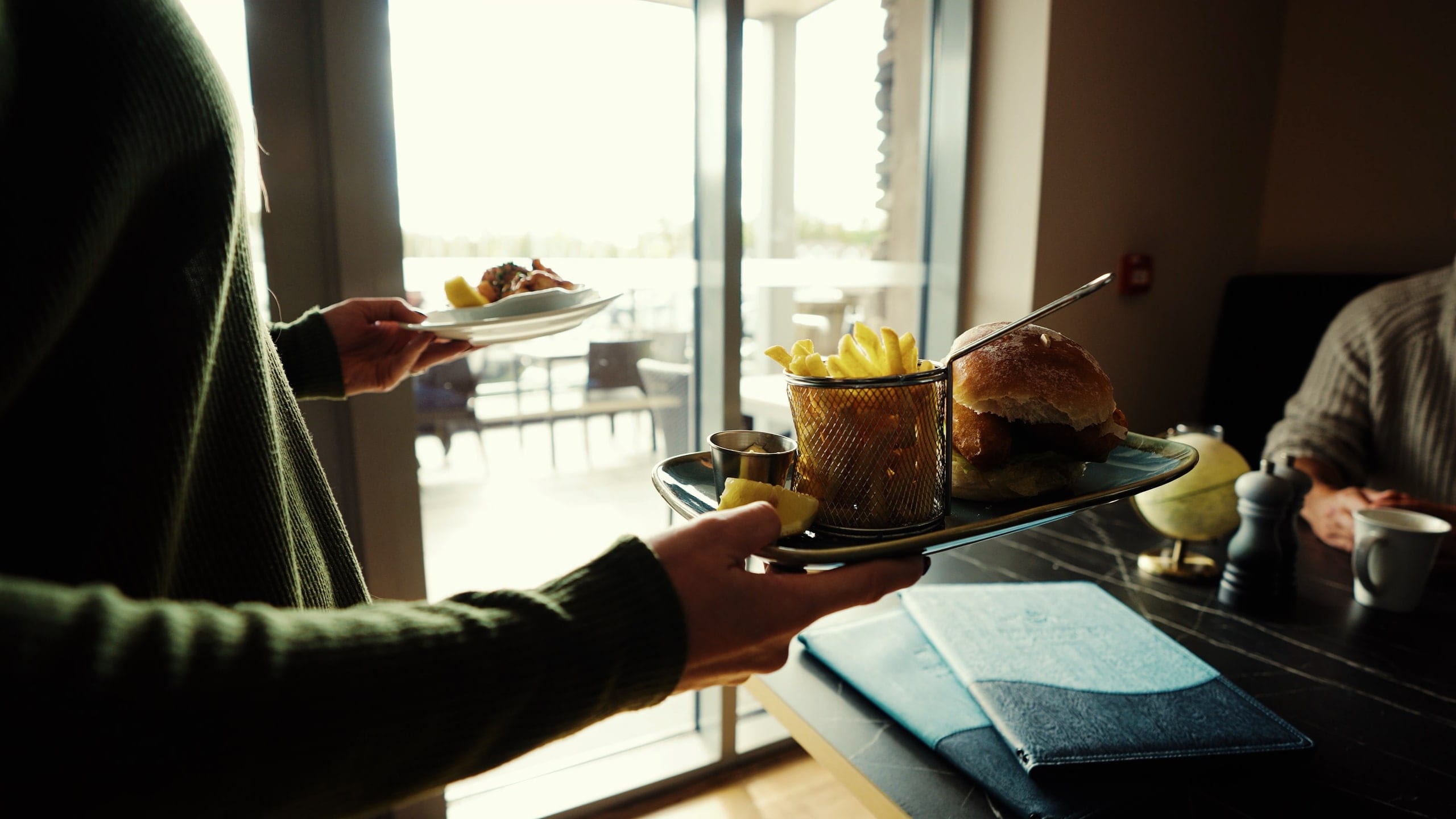 A server carrying plates to a window table in a bright restaurant, a still from a Flexxed hospitality film