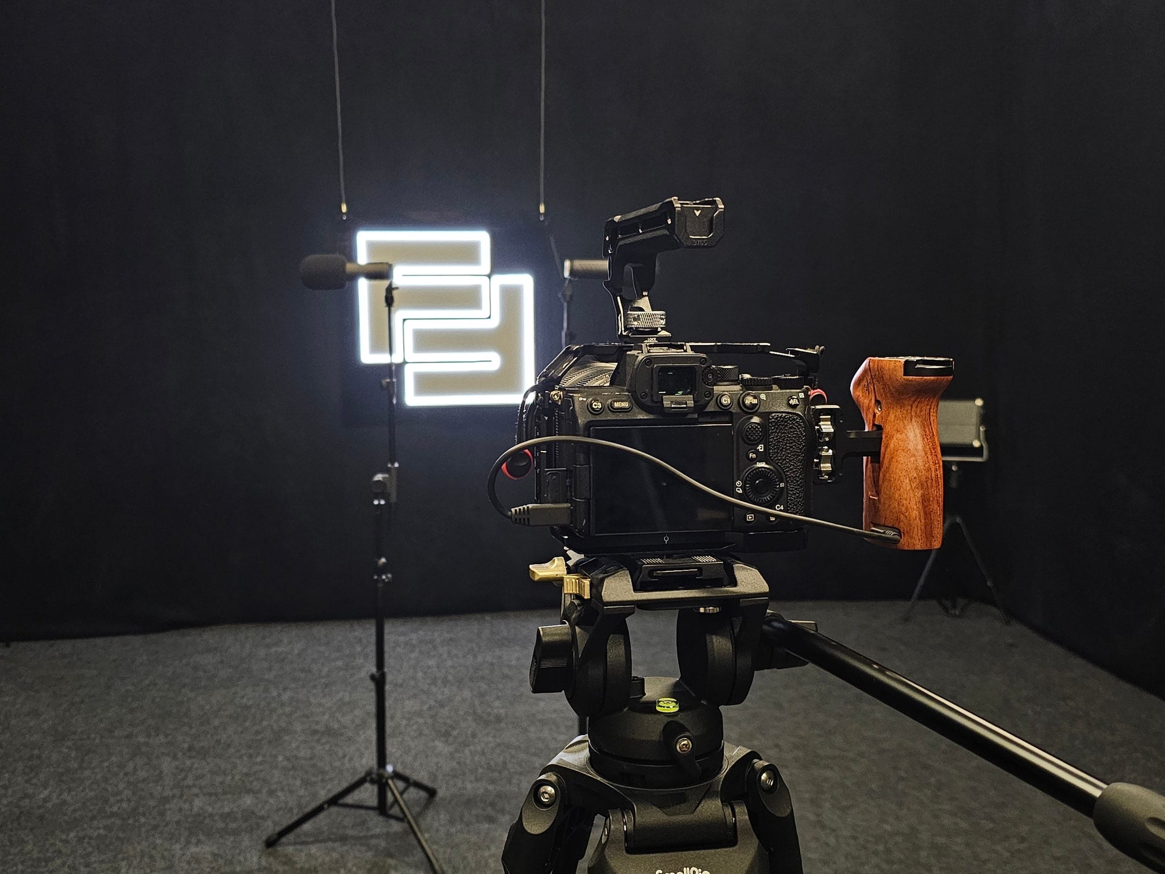 A camera on a tripod in the black studio, framing microphones on stands in front of a lit logo sign