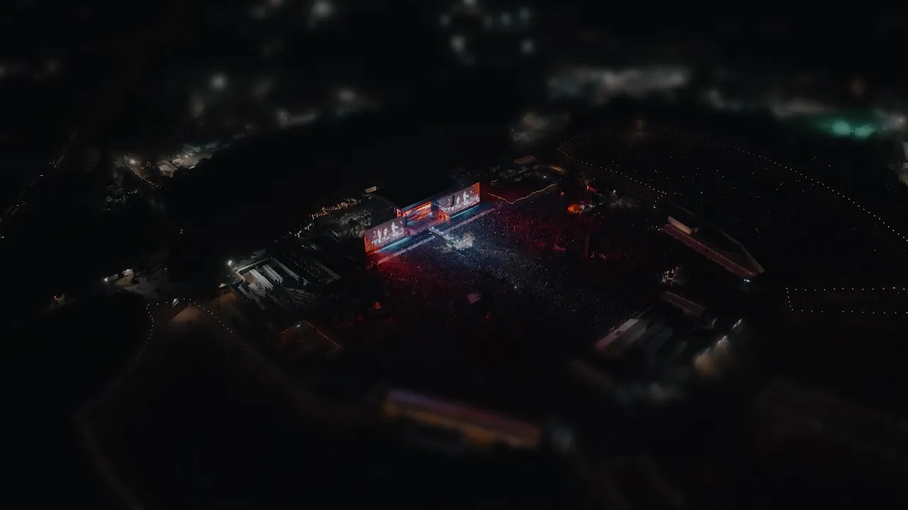 A festival site at night from the air, the stage lit red and white above a packed crowd